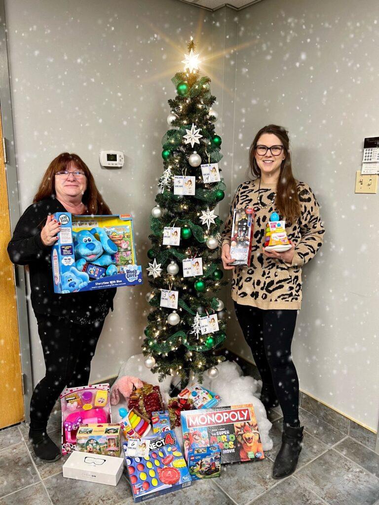 Staff at Cornwall hold toy gifts for donations and stand next to a stack of gifts and a Christmas tree.