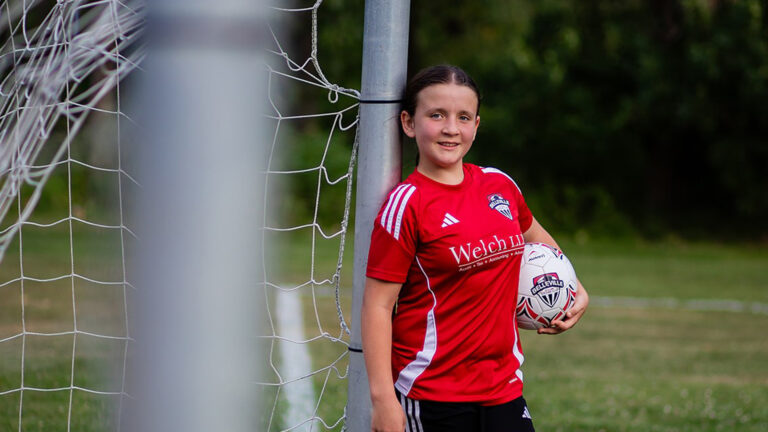 A young girl, a member of the Belleville U11 Girls Soccer Team, leans against a soccer net as she holds a Belleville soccer ball, wearing a red jersey with the Welch LLP logo across the front.