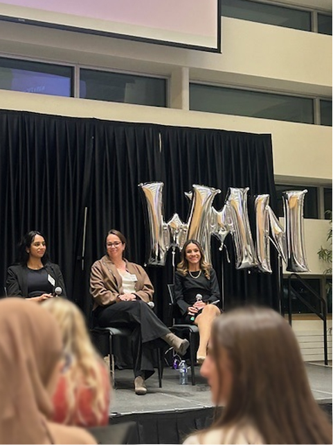 Monica Valbuena sitting on a stage holding a microphone among other panelists with WMN balloons behind her.